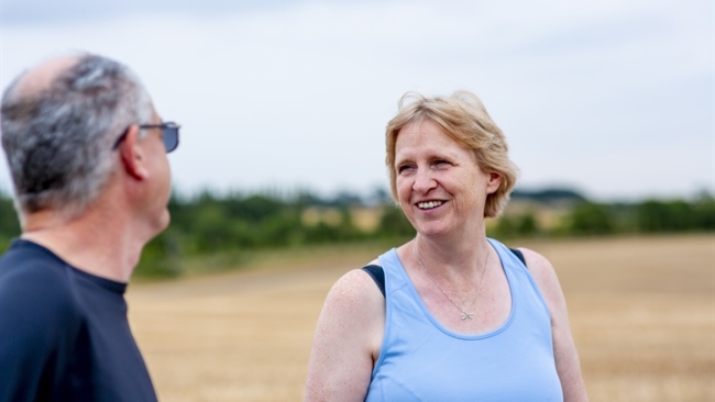 Two people talking outdoors in a sunny, open field; a woman in a light blue tank top smiles at a man wearing dark clothing.