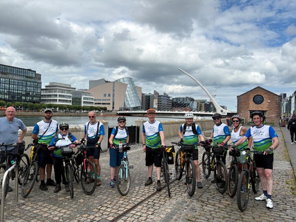 Group of cyclists posing with their bikes along a cobblestone riverside promenade, with modern city buildings and a curved bridge in the background.