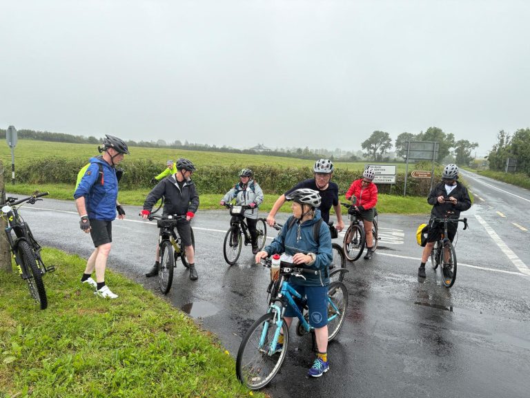 Group of cyclists wearing helmets stop on a wet rural road, chatting beside their bikes as signs point ahead for routes.
