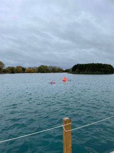 Overcast lake with red buoys marking a course, a wooden post and rope in the foreground, and a tree-covered island in the distance.