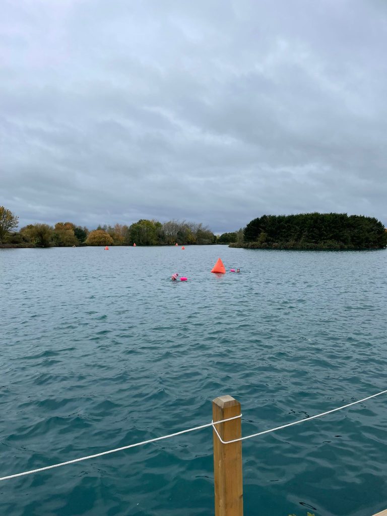 Overcast lake with red buoys marking a course, a wooden post and rope in the foreground, and a tree-covered island in the distance.