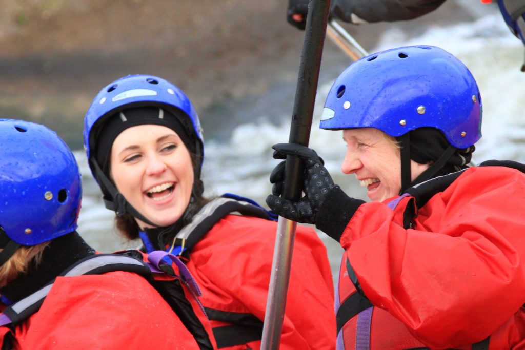 Two women in blue helmets and red life jackets laugh together while holding a paddle on a rafting trip by whitewater.