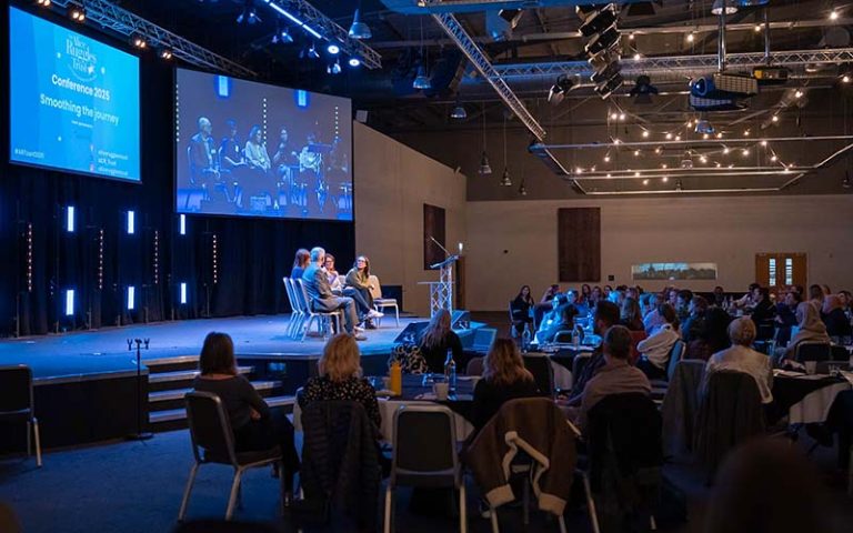 Panel discussion on a stage at a conference, with screens showing the speakers and an audience seated at tables below.