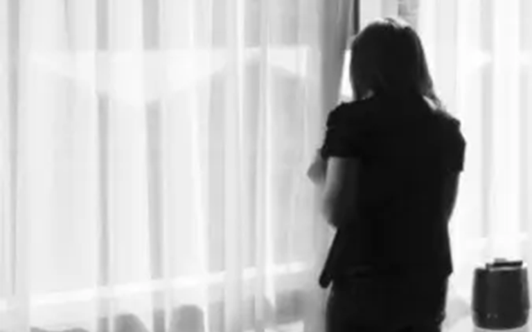 Person stands by a window with sheer curtains, seen from behind in a black-and-white photo.