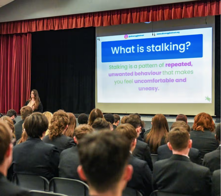 Audience seated in a auditorium watching a presenter beside a projected slide that asks 'What is stalking?'