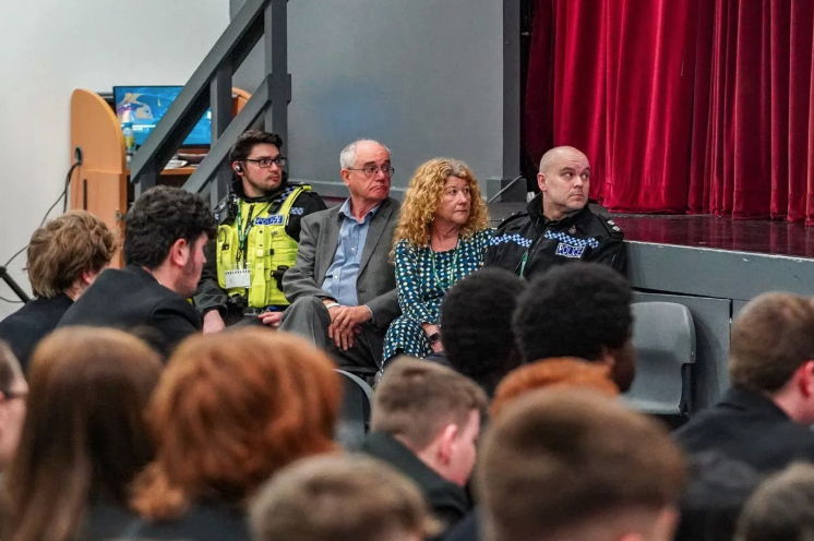 Audience seated in a hall watching a stage; a man in a neon-yellow security jacket and two civilians sit beside a bald police officer near the red-curtained stage.